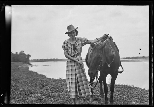 A black-and-white image of a White woman wearing a cowboy hat and plaid dress holding the reins and saddle of the horse next to her.