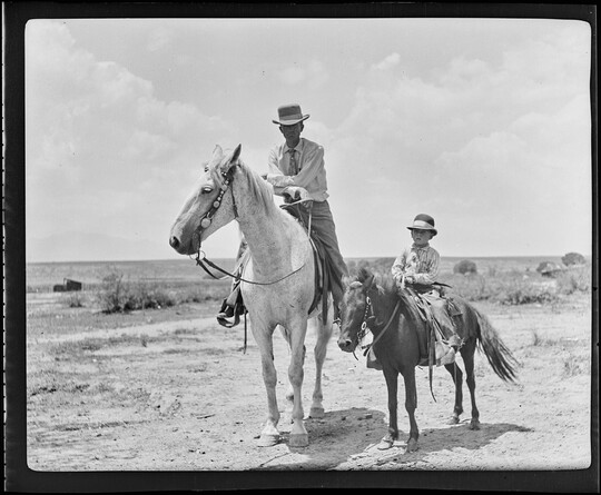 A black-and-white image of a White man wearing a cowboy hat on horseback and, next to him, a White child wearing a cowboy hat on a pony.