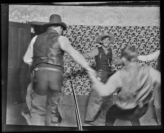 A black-and-white image of White cowboys holding hands dancing in a circle inside a room with bold patterned wallpaper.
