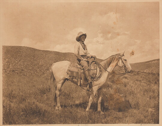 A sepia-toned photograph of a cowboy on a horse standing in a hilly plain.