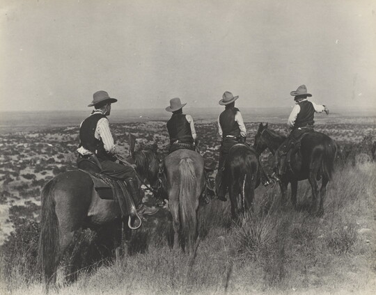 A black-and-white photograph of four cowboys on horseback, viewed from behind, looking over plains in the distance.