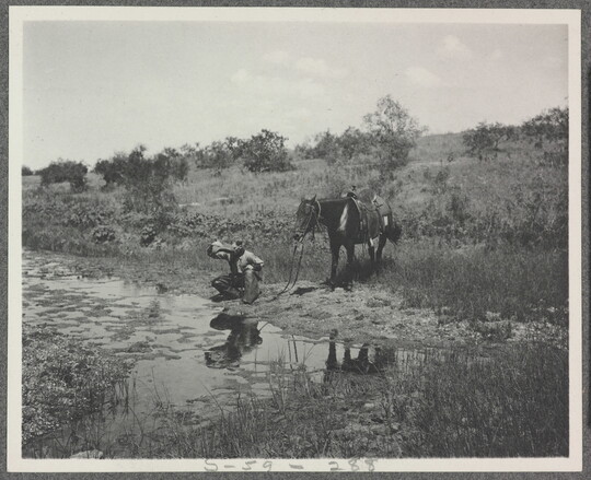 A black-and-white photograph of a cowboy crouched next to a stream drinking water from his hat as his horse stands behind him.
