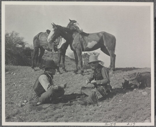 A black-and-white photograph of two cowboys playing a card game on the ground, while their horses stand nearby.