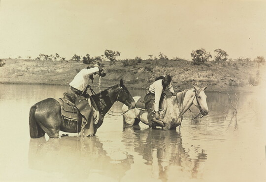 A sepia-toned photograph of two cowboys riding their horses through a river.