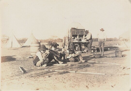 A black-and-white photograph of a group of cowboys, several seated on the ground, one walking, and one preparing food at a wagon; tipis in the background.
