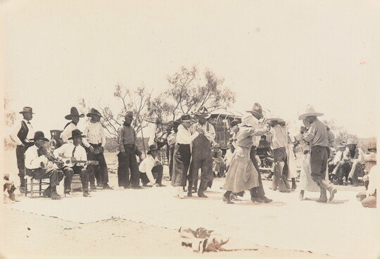 A black-and-white photograph of a group of people outdoors, most wearing cowboy hats, dancing as a band plays.