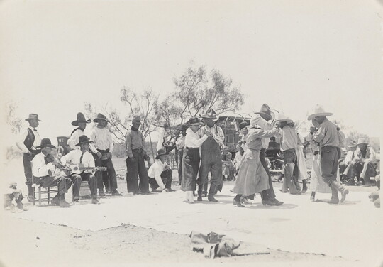 A black-and-white photograph of a group of people outdoors, most wearing cowboy hats, dancing as a band plays.