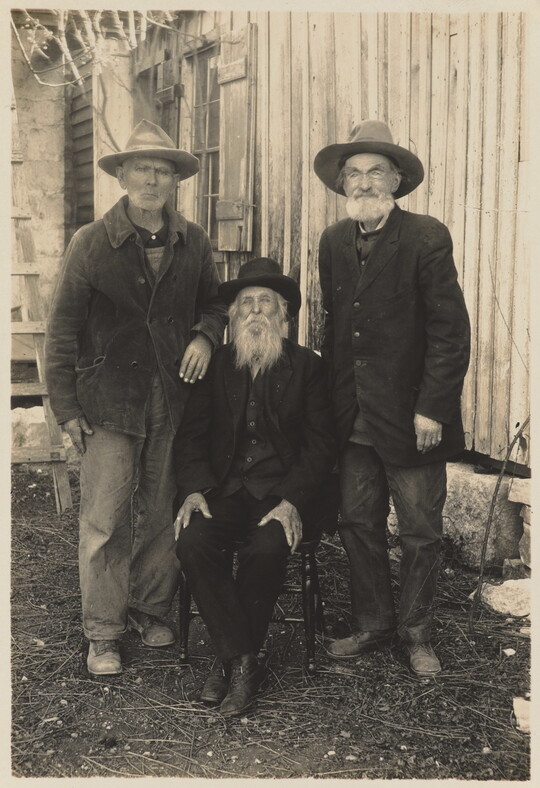 A black-and-white photograph of three men, two standing and one sitting, outside of a wood house, all wearing coats and hats.