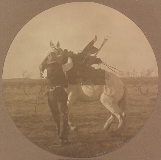 A sepia-toned circular photograph of a cowboy trying to calm a white horse.
