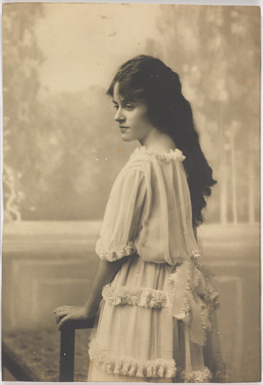A black-and-white photograph of a young White woman with long dark hair wearing a ruffled dress standing back to the camera but head turned in profile.