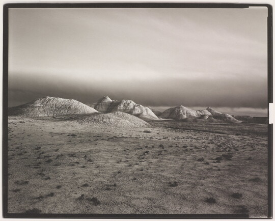 A black-and-white landscape photograph of distant hills at the edge of a flat desert under a dark sky.