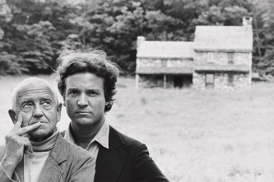 A black-and-white photograph of two White men, one older and one younger, standing in front of a stone farmhouse in the distance.