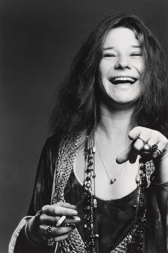 A black-and-white portrait photograph of a White woman with long hair, an open-mouthed smile, holding a cigarette in one hand and pointing at the camera with the other.