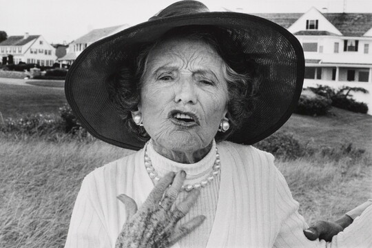 A black-and-white photograph of an elderly White woman wearing a wide-brimmed sunhat, pearl earrings and necklace, looking as if she is speaking.