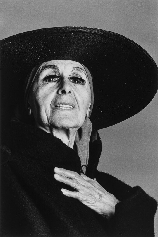 A black-and-white photograph of an older White woman wearing a black hat with a large round brim, excessive eye make-up, black clothing, and her hand over her heart.