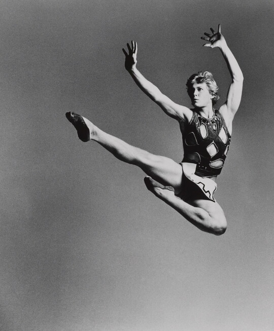 A black-and-white photograph of a White man leaping into the air kicking one leg straight out, the other bent inward, both arms up above his head.