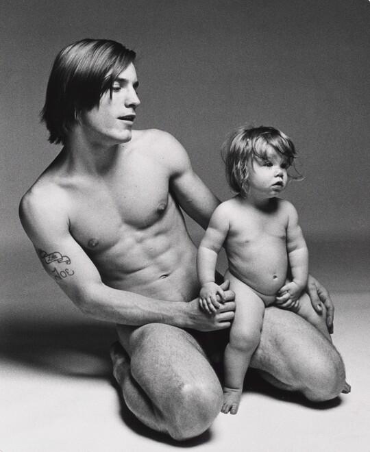 A black-and-white photograph of a nude White man kneeling and holding a nude White toddler on his knee.