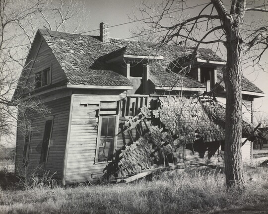 A black-and-white photograph of an abandoned, two-story, wood home that is falling apart.