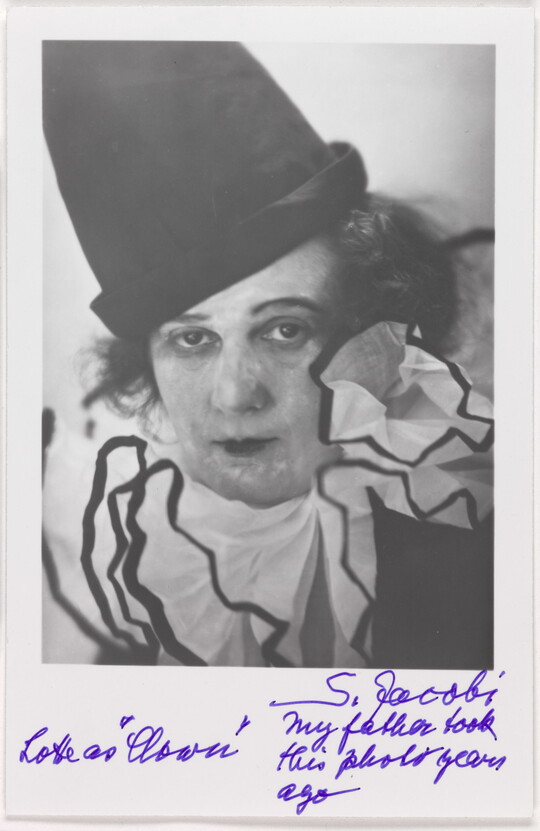 A black-and-white photograph of a woman wearing blotchy white face paint, a tall black hat, and frilly white collar, above a handwritten note.