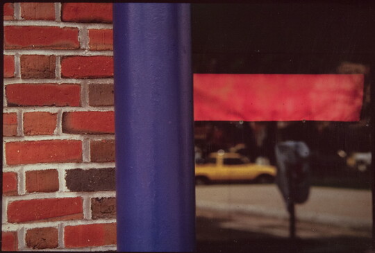 A color photograph of (L-R) a partial red brick wall, a blue pole, glass with red tape on it reflecting a yellow taxi and a parking meter.
