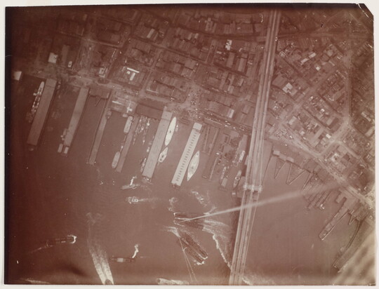 A black-and-white photograph of an aerial view of a boat harbor next to a city and a long bridge crossing the water into the city.