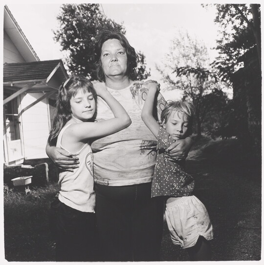 A black-and-white photograph of a middle-aged White woman with her arms around two young children who are hanging on her shoulders.