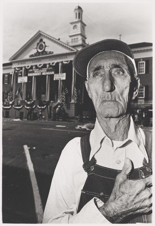 A black-and-white photograph of an elderly White man wearing a baseball cap, overalls, hand over his heart standing in front of a courthouse decorated in flags and buntings.