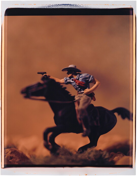 A color photograph of a White cowboy on a galloping horse, in profile, pointing a revolver in front of him.
