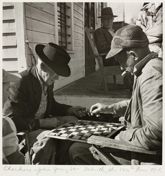 A black-and-white photograph of two older White men playing checkers and smoking cigars in front of a building as another man watches in the background.