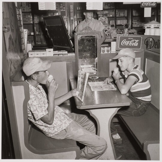 A black-and-white photograph of two White boys wearing baseball hats and eating ice cream cones in a booth.