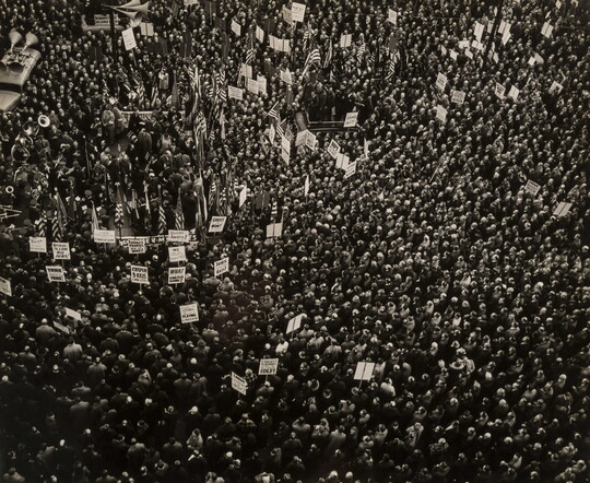 A black-and-white aerial photograph of a crowd of people, some carrying signs and American flags.