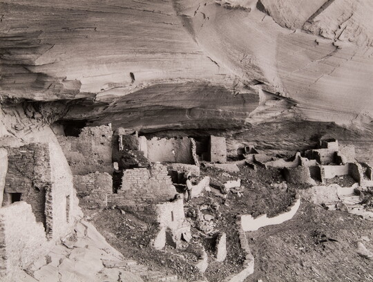 A black-and-white photograph of adobe ruins under an overhanging cliff ledge.