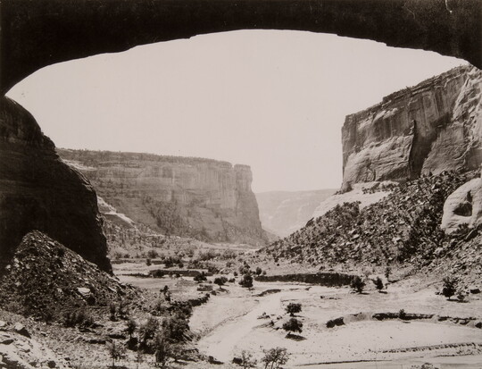 A black-and-white photograph of a canyon landscape framed by an arched rock overhang.