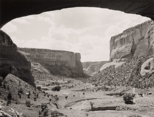 A black-and-white photograph of a canyon landscape framed by an arched rock overhang.