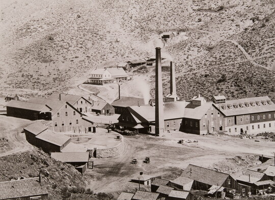 A black-and-white photograph of industrial buildings with smoke stacks in a rocky valley.
