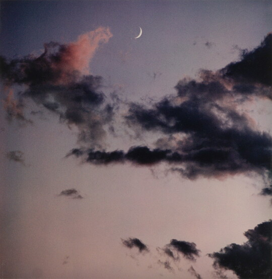A color photograph of a blue-gray sky, dark clouds, and a crescent moon.
