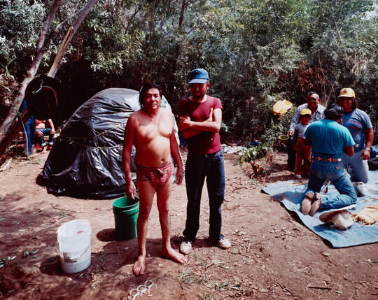 A color photograph of two medium-skinned men: one man wearing a loin cloth and the other in jeans and a t-shirt standing in a wooded setting.