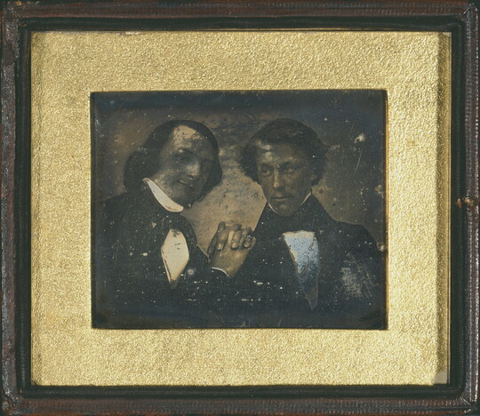 A very old, worn black-and-white photograph in a gold frame of two men in formal attire posing for the camera.