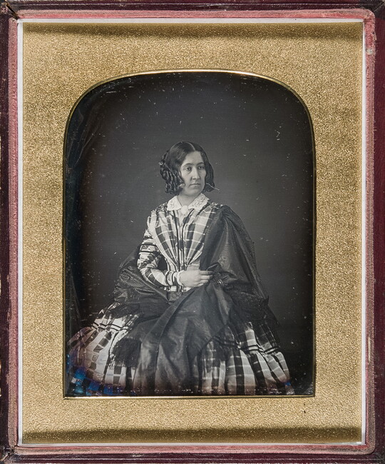 A black-and-white portrait photograph in a gold-frame of a White woman with her hair styled in ringlets, wearing a plaid dress with a full skirt and a shawl wrapped around her shoulders.