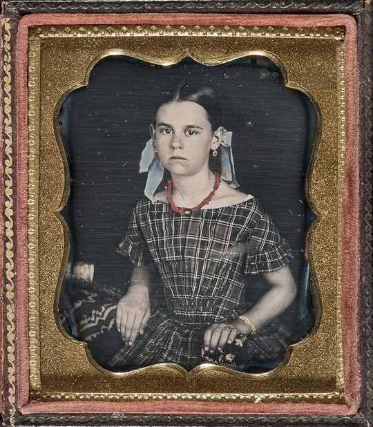 A hand-colored photograph in a decorative gold frame of a young White girl seated, wearing a plaid dress and bows in her hair.