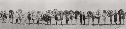 A black-and-white panoramic photograph of a group of women, lined side-by-side on a beach, wearing elaborate bathing costumes, hats, and holding parasols.