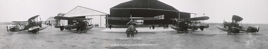A black-and-white panoramic photograph of an airplane hanger with five biplanes in the foreground.