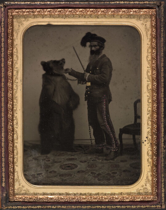 A black-and white-photograph in a decorative gold frame of a bearded man next to a standing bear that has a chain around its neck.