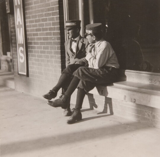 A black-and-white photograph of two young White men, both in military-style caps, jackets, ties, knickers, and hose, sitting on a window ledge.