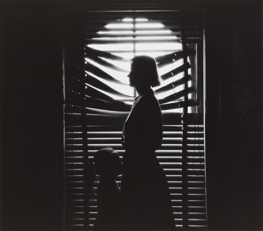 A black-and-white photograph of the silhouettes of a woman and young girl facing each other in front of a window with light coming through partially closed blinds.