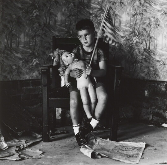 A black-and-white photograph of a young White boy in a chair holding a naked doll on his lap and waving an American flag.