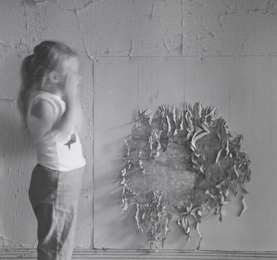 A black-and-white photograph of a young White girl in profile, hair in a pony tail, one arm blurred with motion, in front of a wall of peeling paint.