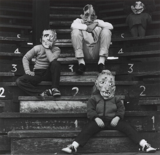 A black-and-white photograph of four children in grotesque costume masks sitting on wooden steps.