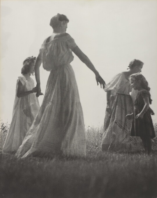 A black-and-white photograph of White women and children wearing long dresses holding hands to form a circle in a grassy field.