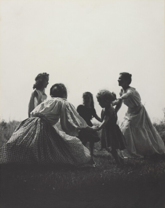 A black-and-white photograph of White women and children wearing long dresses holding hands to form a circle in a grassy field.